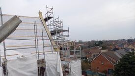 L’image montre un site de construction où un bâtiment est entouré d’échafaudages, indiquant des travaux de rénovation ou de construction. Des bâches de protection sont visibles sur certaines parties de la structure. À l’arrière-plan, on aperçoit un quartier résidentiel avec des maisons et des arbres qui s’étendent au loin. Le ciel est couvert, suggérant une journée nuageuse.