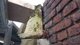 L’image représente un coin architectural où se rencontrent deux types de matériaux : à droite, un mur en briques rouges bien alignées, et à gauche, des pierres de taille irrégulières, recouvertes en partie de mousse verte. Au-dessus de cette jonction murale, une structure composée de bois et de métal est visible, suggérant un élément de charpente ou de support.
