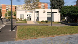 L’image montre un petit bâtiment moderne à façade blanche, doté de grandes fenêtres. Une enseigne au-dessus de l’entrée indique "Pole Social". Un chemin pavé mène à l’entrée, bordé de pelouse et de quelques arbustes. À l’arrière-plan, on aperçoit des immeubles d’habitation plus grands et des arbres. L’environnement semble urbain, avec des trottoirs aménagés et des lampadaires visibles au premier plan.