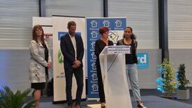 Quatre personnes se tiennent sur une scène, dont Madame La Maire derrière un pupitre portant l’inscription « COMPLEXE SPORTIF JEAN-PHILIPPE GATIEN ». En arrière-plan, des banderoles affichent des logos et des textes, notamment « Hauts-de-France » et « Ville de Lille – Une volonté partagée ». L’événement semble se dérouler en intérieur, dans un cadre officiel ou communautaire, possiblement lié au sport. Des plantes décoratives sont disposées autour de la scène.
