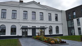 L’image montre un grand bâtiment de deux étages avec une façade blanche et de nombreuses fenêtres. L’entrée principale est composée de doubles portes vitrées surmontées d’un petit auvent. Devant le bâtiment, un espace paysager est aménagé avec diverses plantes et fleurs. Le bâtiment semble faire partie d’un ensemble plus vaste, d’autres structures étant visibles de part et d’autre.