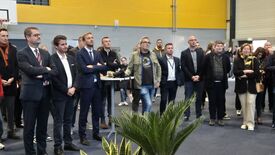 Un groupe de personnes se tient dans un espace intérieur, probablement lors d’un événement ou d’un rassemblement. En arrière-plan, on distingue un mur jaune et gris, un panier de basket, ainsi que quelques plantes au premier plan. Les personnes présentes portent des tenues variées, allant du costume à des vêtements plus décontractés, et certaines portent des lanyards. Leurs visages sont floutés pour préserver leur anonymat.