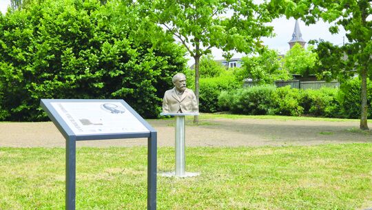 L’image montre une statue en buste posée sur un piédestal, située dans un espace extérieur paysager. À côté de la statue, un panneau d’information est installé sur un support métallique.

L’environnement comprend des arbres, des buissons, et en arrière-plan, on aperçoit un bâtiment avec un clocher ou une flèche, suggérant une église ou un édifice historique.