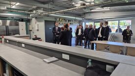 Un groupe de personnes se tient dans ce qui semble être un établissement moderne à vocation industrielle ou éducative. Le plafond présente des conduits et tuyaux apparents. Le long d’un mur, plusieurs trophées sont exposés sur des étagères. Au premier plan, un long comptoir est équipé de prises électriques et d’un carnet. À l’arrière-plan, des tables et des chaises sont disposées, et de grandes fenêtres laissent entrer la lumière naturelle. Les personnes présentes portent des tenues formelles, ce qui suggère une visite officielle ou une inspection du lieu.
