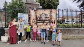 Un groupe de personnes, adultes et enfants, se tient devant une banderole portant l’inscription « BRUAY-LANTA ». La banderole est décorée d’un motif tribal avec des flammes de chaque côté. Les participants tiennent des certificats ou des récompenses. En arrière-plan, un panneau d’affichage présente plusieurs affiches et annonces.