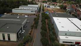 L’image montre une vue aérienne d’une zone industrielle comportant plusieurs grands bâtiments, des parkings et une route bordée d’arbres traversant le centre de l’image. De nombreux véhicules sont stationnés à proximité des bâtiments. L’environnement alentour comprend des habitations résidentielles et des espaces verts. L’un des bâtiments porte une enseigne indiquant "POLE ARTISANAL BRUAYCO".