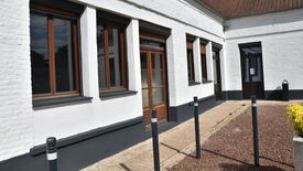 Façade extérieure d’un bâtiment en briques blanches, comportant plusieurs fenêtres et portes aux encadrements en bois. Des bornes noires sont alignées le long du trottoir devant le bâtiment. Sur la droite, un petit espace paysager contient quelques plantes.