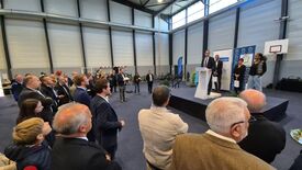 Un groupe de personnes est rassemblé dans un grand espace intérieur, probablement un gymnase ou une salle de conférence. Une scène est installée, avec plusieurs personnes debout, dont une prenant la parole à un pupitre. Le public, debout, fait face à la scène et écoute l’intervenant. Des banderoles et des panneaux sont visibles derrière le pupitre, accompagnés de plantes décoratives. La salle est dotée de hauts plafonds et de grandes fenêtres laissant entrer la lumière naturelle. Un panier de basket est visible sur la droite de l’image.