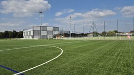 Un terrain de football en gazon synthétique bien entretenu est visible, avec des lignes blanches et bleues délimitant les zones de jeu. À l’arrière-plan, un grand bâtiment à la façade grise et blanche, probablement un gymnase ou un complexe sportif, domine la scène. Plusieurs mâts d’éclairage entourent le terrain, indiquant qu’il peut être utilisé en soirée. Le ciel est dégagé avec quelques nuages épars, suggérant une météo favorable. Aucune personne n’est visible sur l’image.