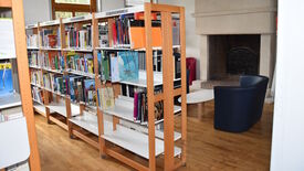 L’image montre une section d’une bibliothèque. Plusieurs étagères sont remplies de livres, avec des étiquettes indiquant les catégories telles que "Géographie", "Histoire de l'Europe" et "Inde". À l’arrière-plan, un coin lecture est aménagé avec deux fauteuils, une petite table et une cheminée, créant une ambiance confortable et propice à la lecture.