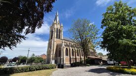 L’image représente une église en briques rouges dotée d’un clocher imposant avec une horloge visible. L’édifice est entouré de végétation, notamment des arbres et des buissons. Le ciel est partiellement couvert de nuages, laissant apparaître quelques zones de ciel bleu. À l’arrière-plan, on distingue des bâtiments résidentiels. Une clôture métallique est visible sur la gauche. Au premier plan, un chemin en gravier mène à l’église, et plusieurs véhicules sont stationnés sur la droite.