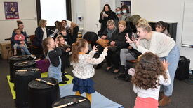 L'image montre une scène d'activité en intérieur. Au premier plan, plusieurs enfants sont debout sur des tapis colorés, effectuant des gestes avec leurs mains. Une femme adulte les accompagne et mime les mêmes gestes. Devant eux, de grands seaux noirs sont disposés, chacun avec des baguettes posées dessus, suggérant une activité de percussion ou de rythme. À l’arrière-plan, plusieurs adultes sont assis sur des chaises, certains prenant des photos ou vidéos avec leurs téléphones.